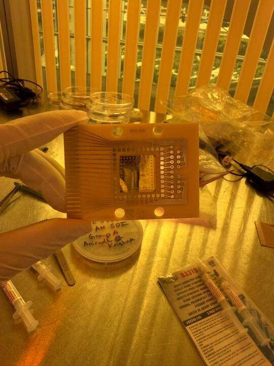 SOI die mounted in the center of a gold-plated carrier board, held in a gloved hand under cleanroom yellow light